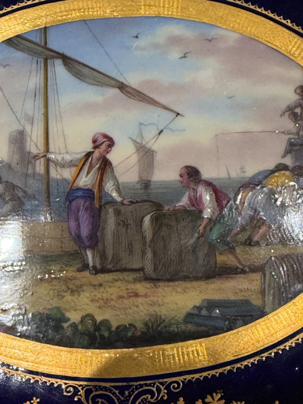 An oval French Sevres porcelain dish featuring a deep cobalt blue rim decorated with intricate gold leaf gilding surrounding a central hand-painted maritime harbour scene. View V