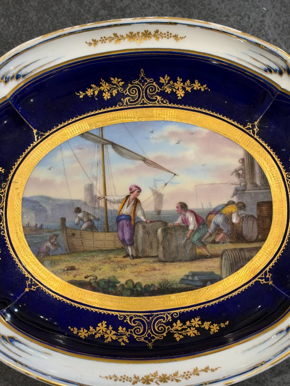 An oval French Sevres porcelain dish featuring a deep cobalt blue rim decorated with intricate gold leaf gilding surrounding a central hand-painted maritime harbour scene. View VI