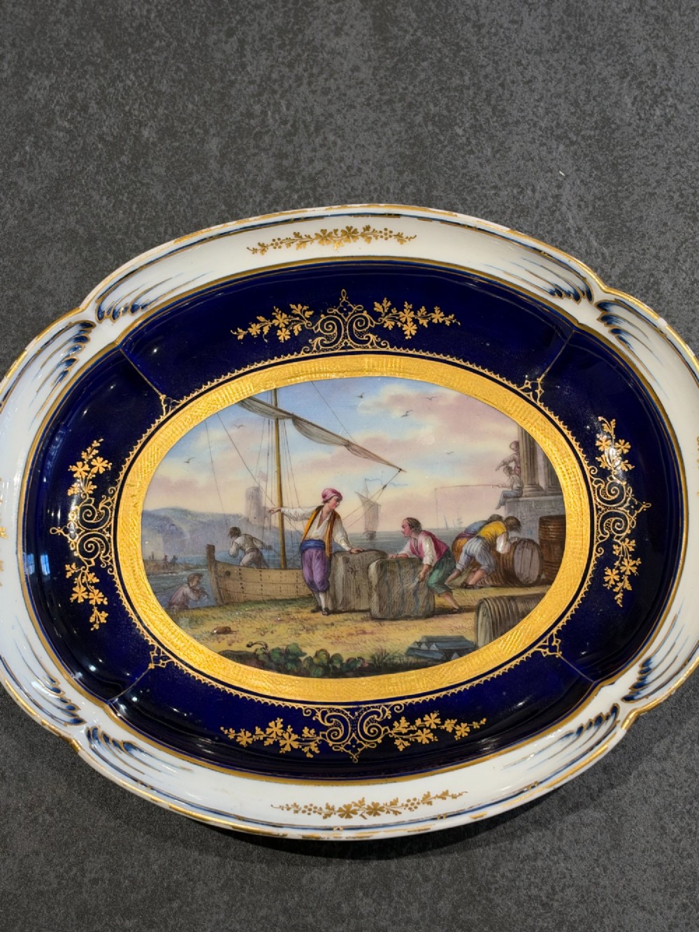 An oval French Sevres porcelain dish featuring a deep cobalt blue rim decorated with intricate gold leaf gilding surrounding a central hand-painted maritime harbour scene. View VII