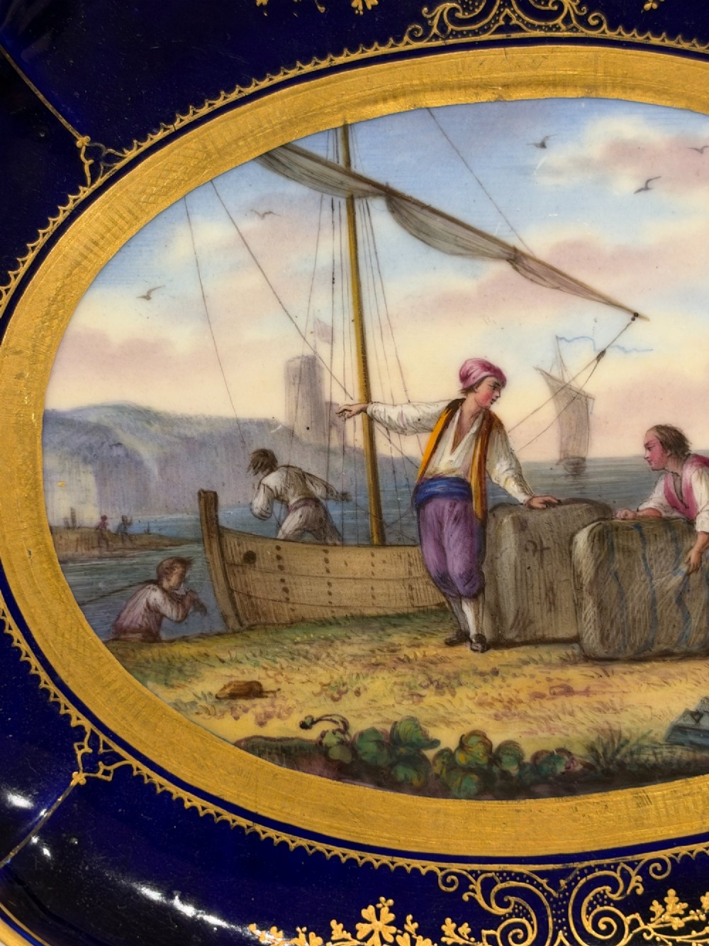 An oval French Sevres porcelain dish featuring a deep cobalt blue rim decorated with intricate gold leaf gilding surrounding a central hand-painted maritime harbour scene. View VIII
