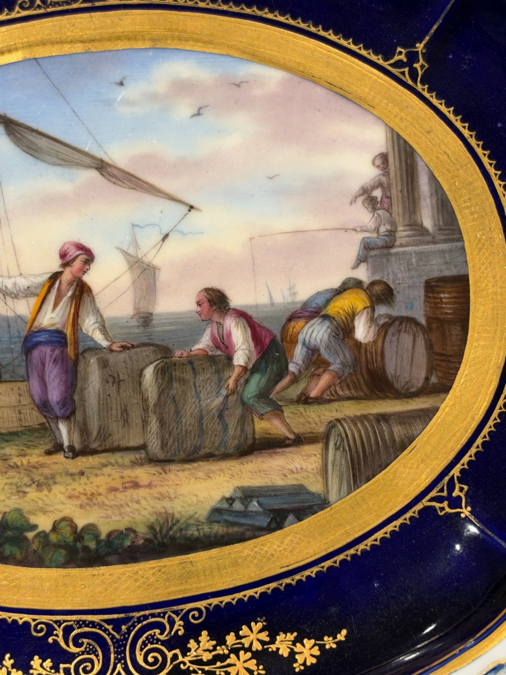 An oval French Sevres porcelain dish featuring a deep cobalt blue rim decorated with intricate gold leaf gilding surrounding a central hand-painted maritime harbour scene. View IX