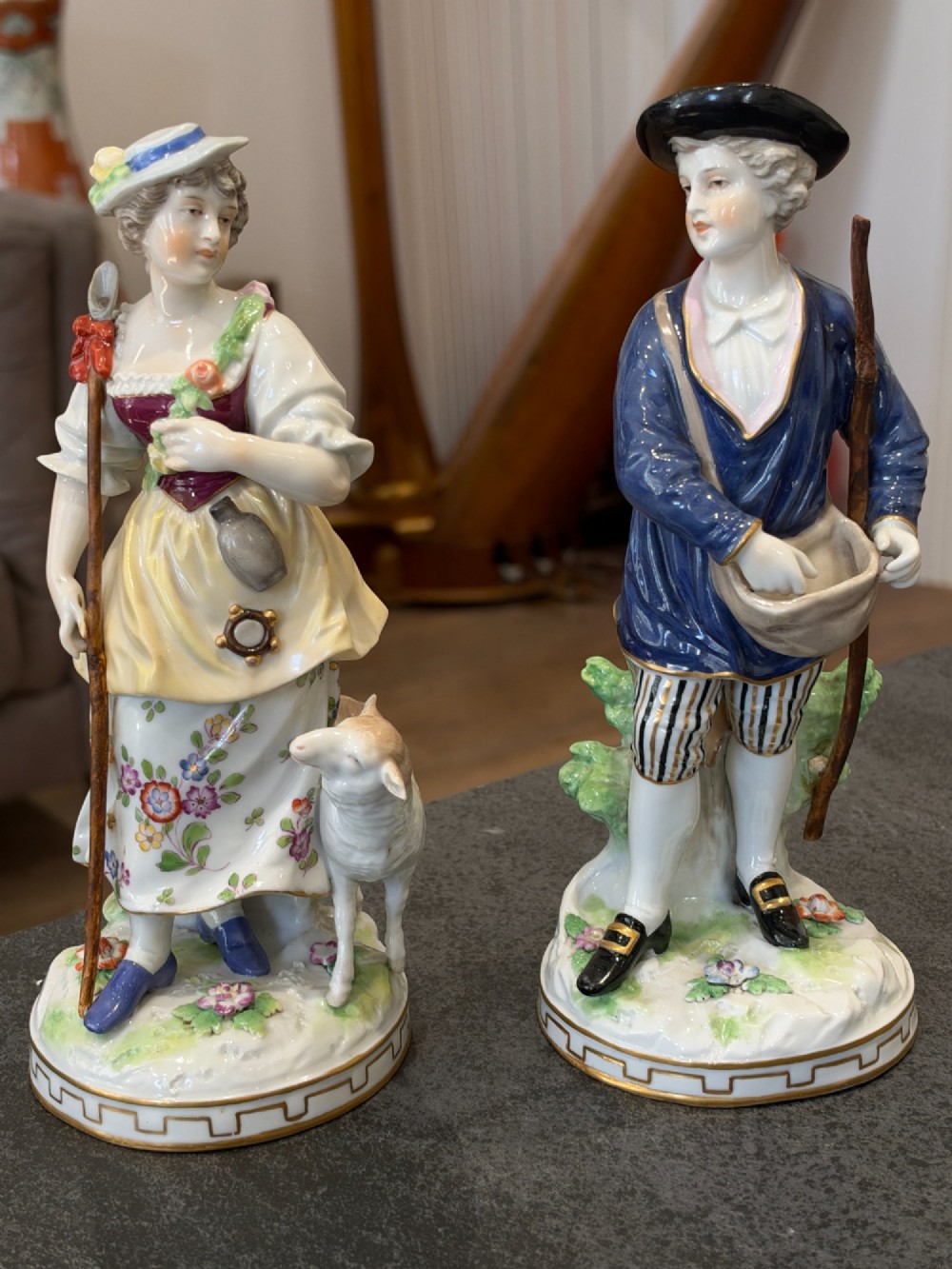 A pair of antique hand-painted German porcelain figures depicting a shepherdess with a lamb and a shepherd holding a basket, set on circular gilded bases.