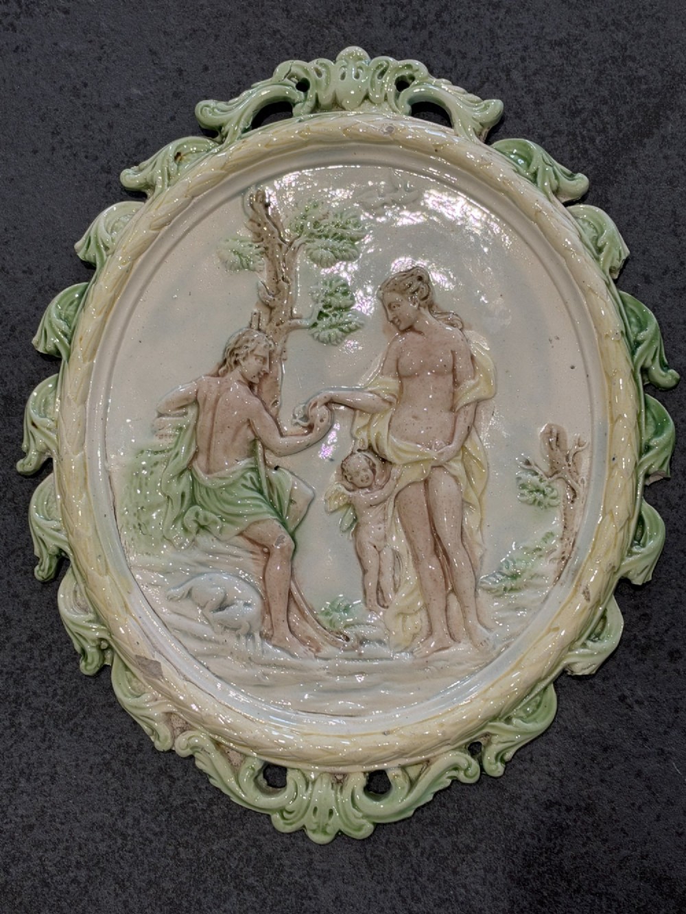 An oval cream and green lead-glazed pearlware plaque featuring relief moulding of classical figures surrounded by an ornate pierced leaf border.