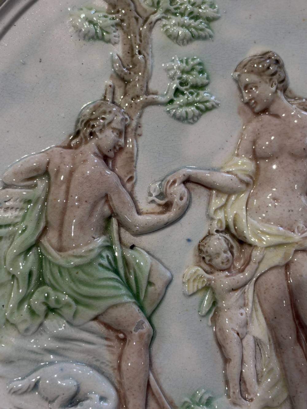 An oval cream and green lead-glazed pearlware plaque featuring relief moulding of classical figures surrounded by an ornate pierced leaf border. View II