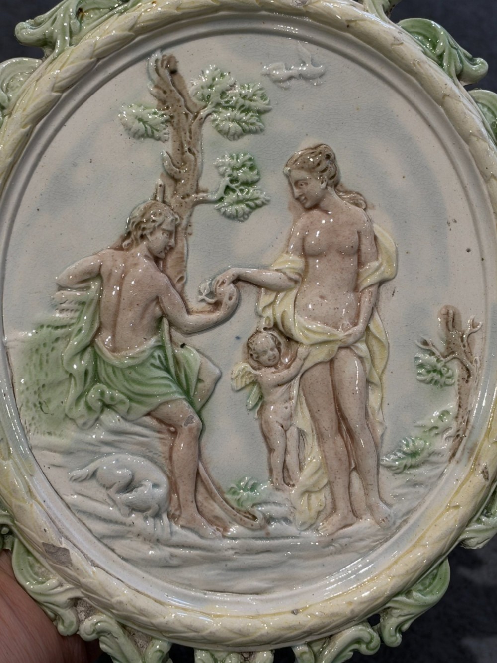 An oval cream and green lead-glazed pearlware plaque featuring relief moulding of classical figures surrounded by an ornate pierced leaf border. View III