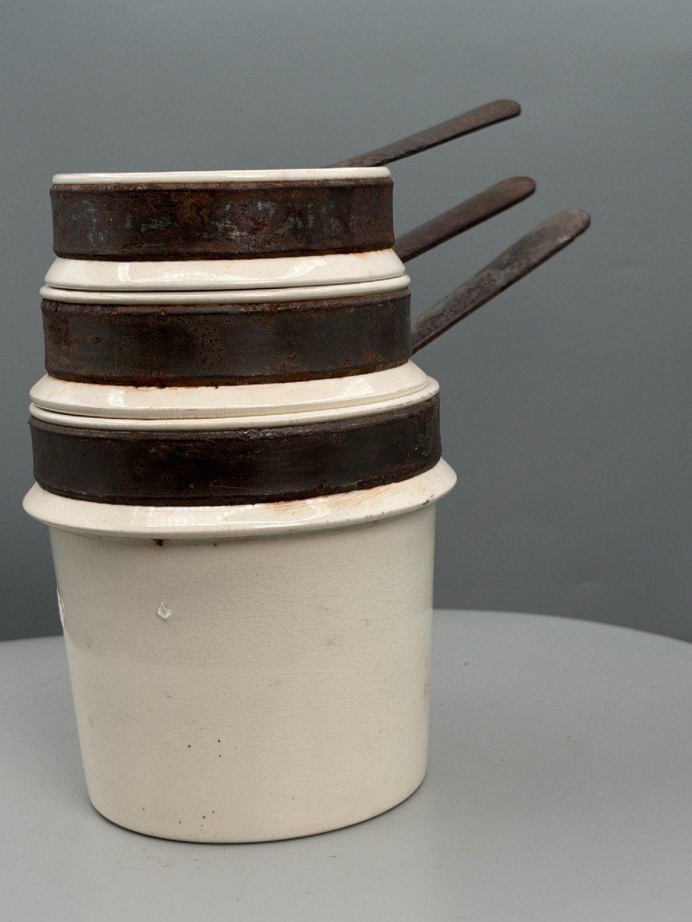 A set of three graduated off-white ceramic cylindrical vessels featuring dark iron bands around the upper rims and extended iron handles. View II