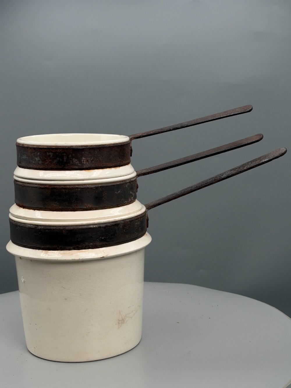 A set of three graduated off-white ceramic cylindrical vessels featuring dark iron bands around the upper rims and extended iron handles. View III