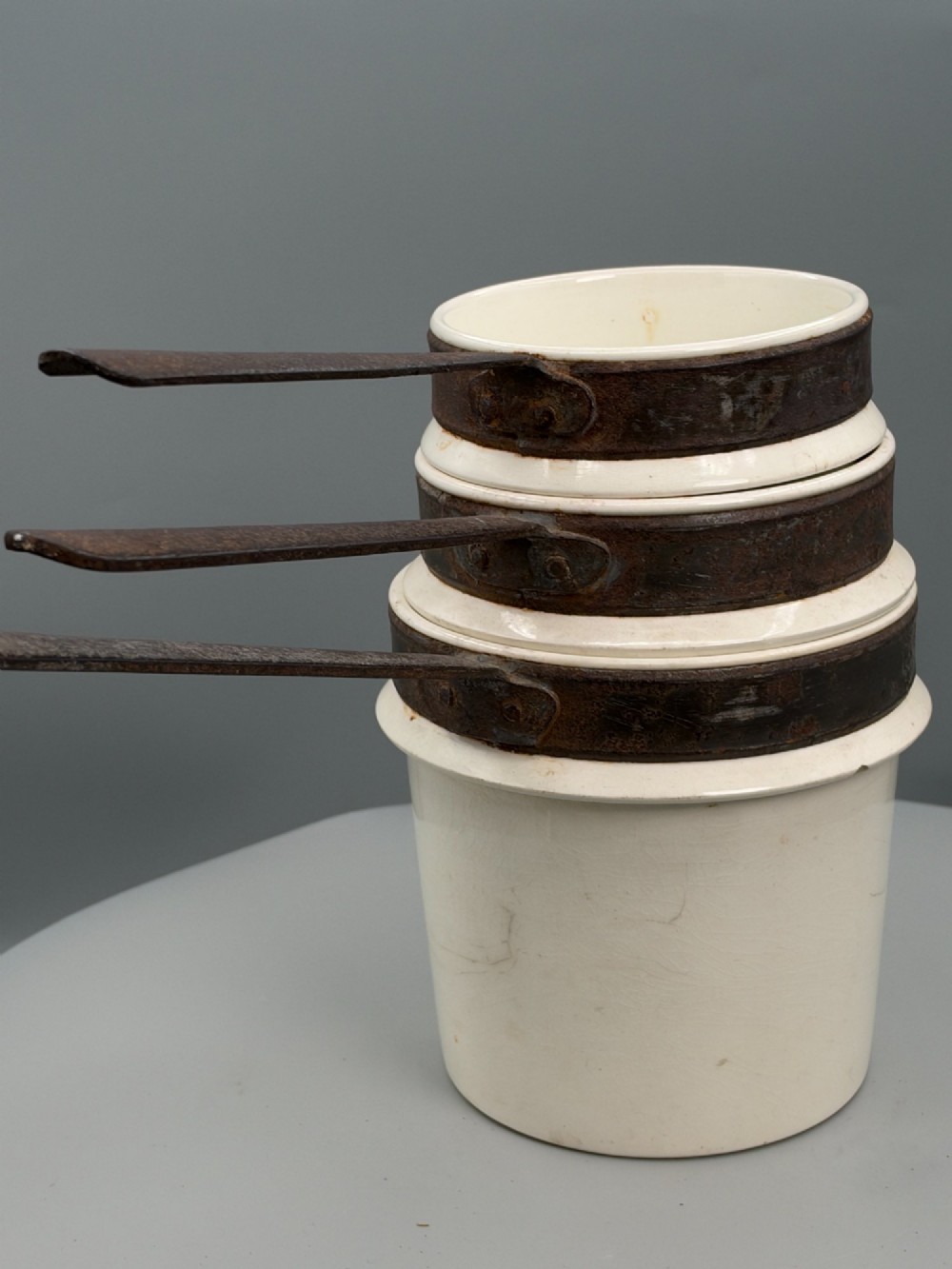 A set of three graduated off-white ceramic cylindrical vessels featuring dark iron bands around the upper rims and extended iron handles. View IV