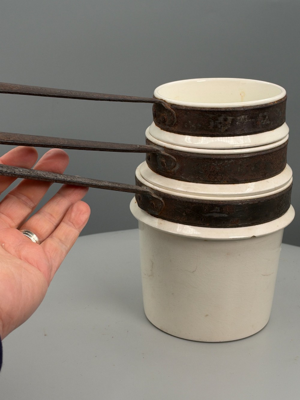 A set of three graduated off-white ceramic cylindrical vessels featuring dark iron bands around the upper rims and extended iron handles. View V