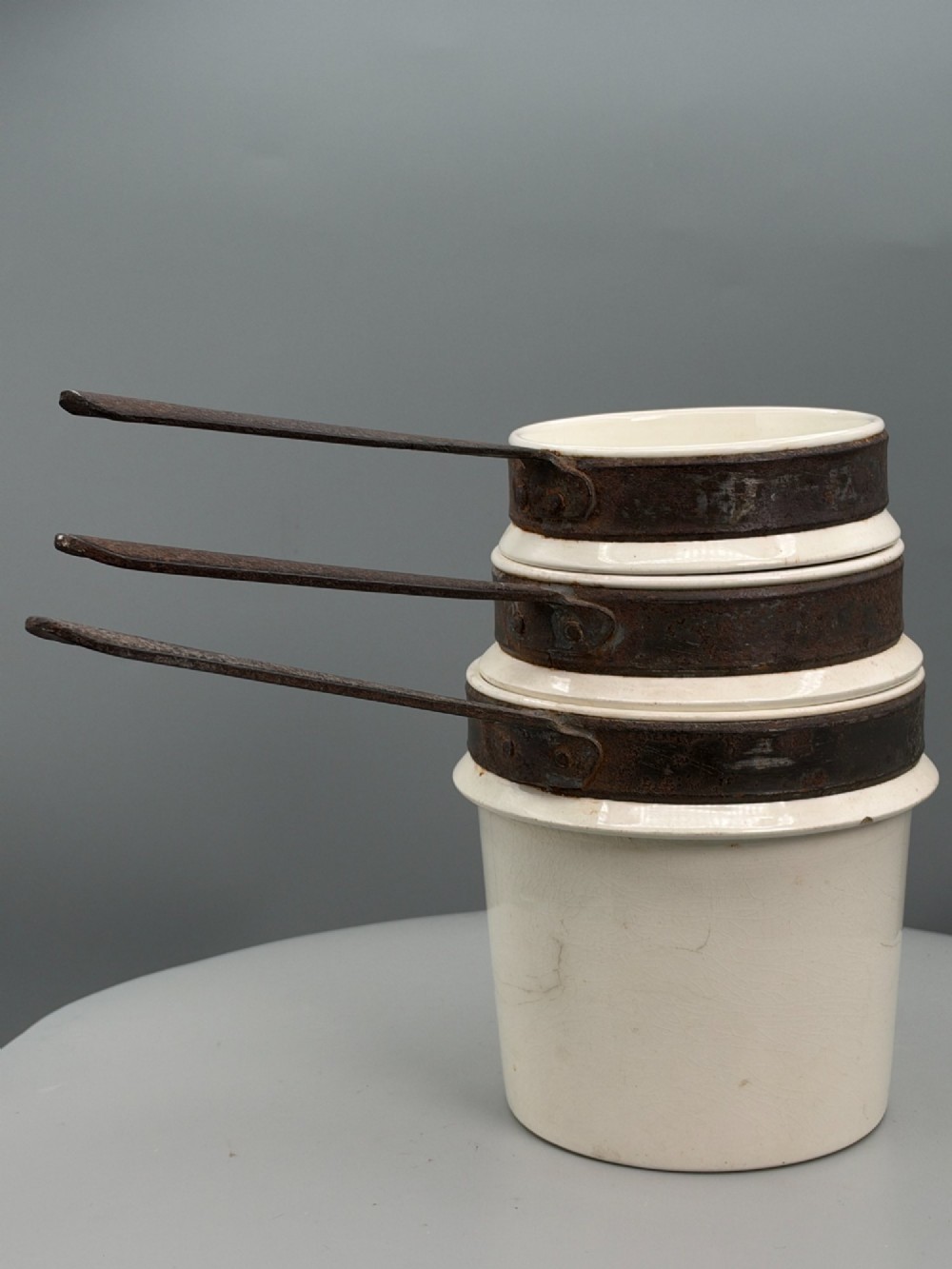A set of three graduated off-white ceramic cylindrical vessels featuring dark iron bands around the upper rims and extended iron handles. View VI