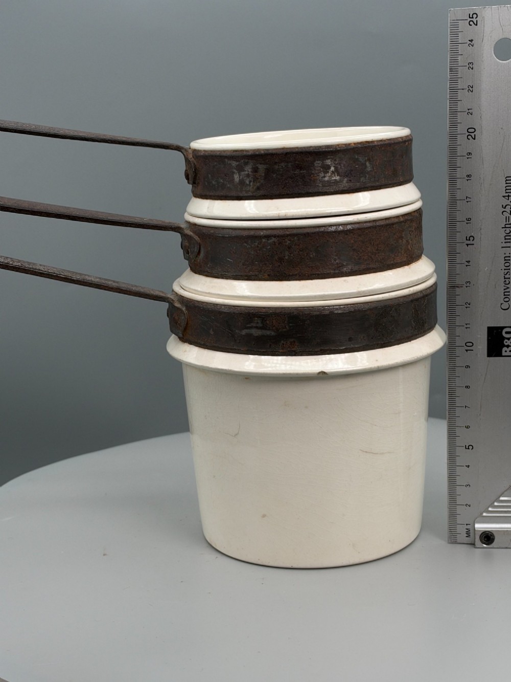 A set of three graduated off-white ceramic cylindrical vessels featuring dark iron bands around the upper rims and extended iron handles. View VII