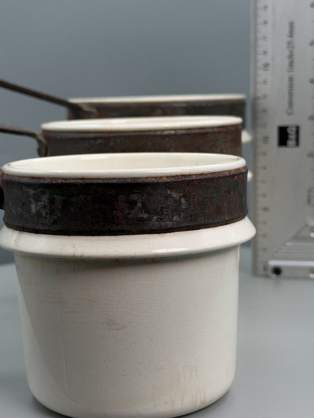 A set of three graduated off-white ceramic cylindrical vessels featuring dark iron bands around the upper rims and extended iron handles. View VIII