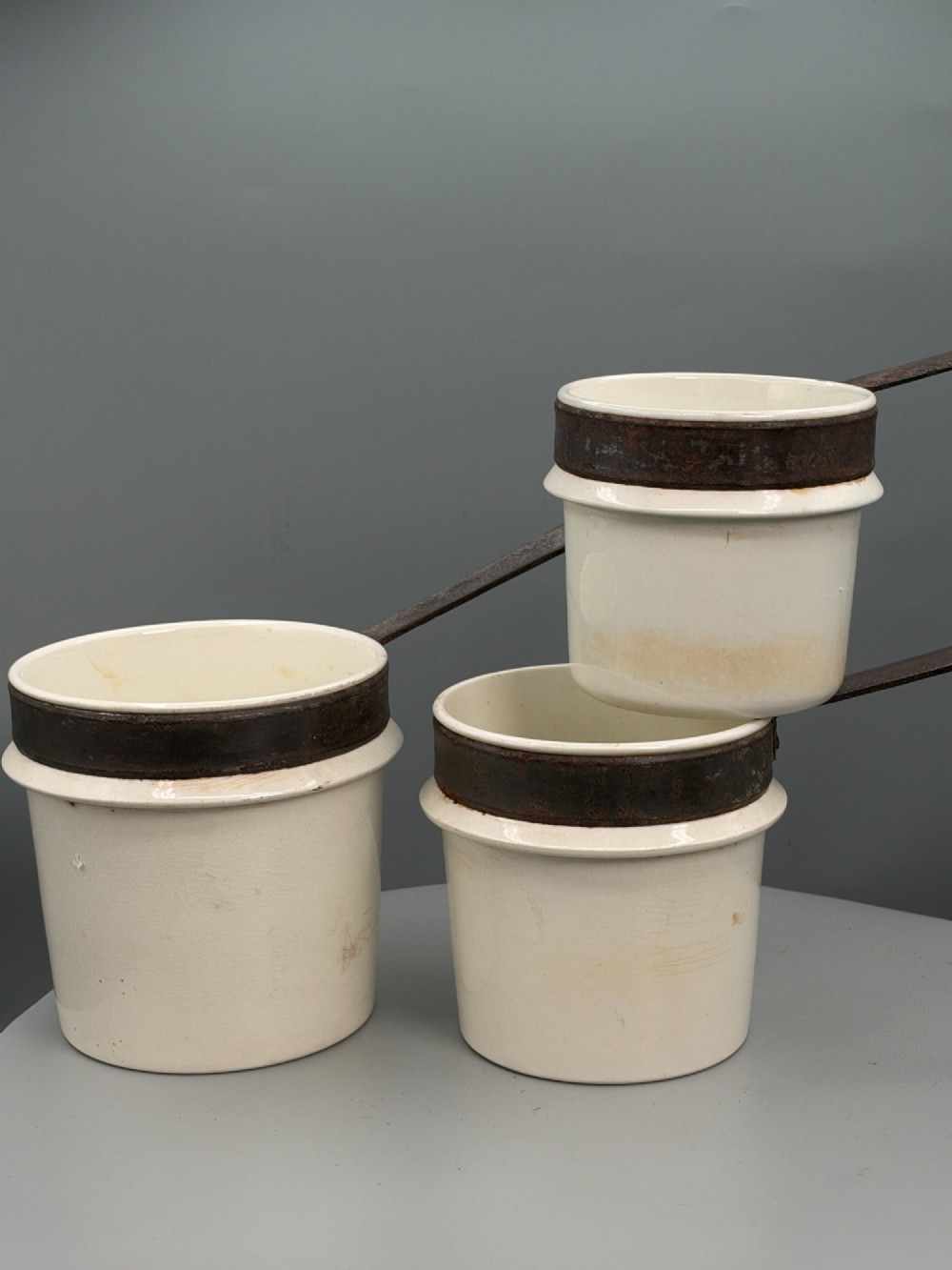 A set of three graduated off-white ceramic cylindrical vessels featuring dark iron bands around the upper rims and extended iron handles. View XII