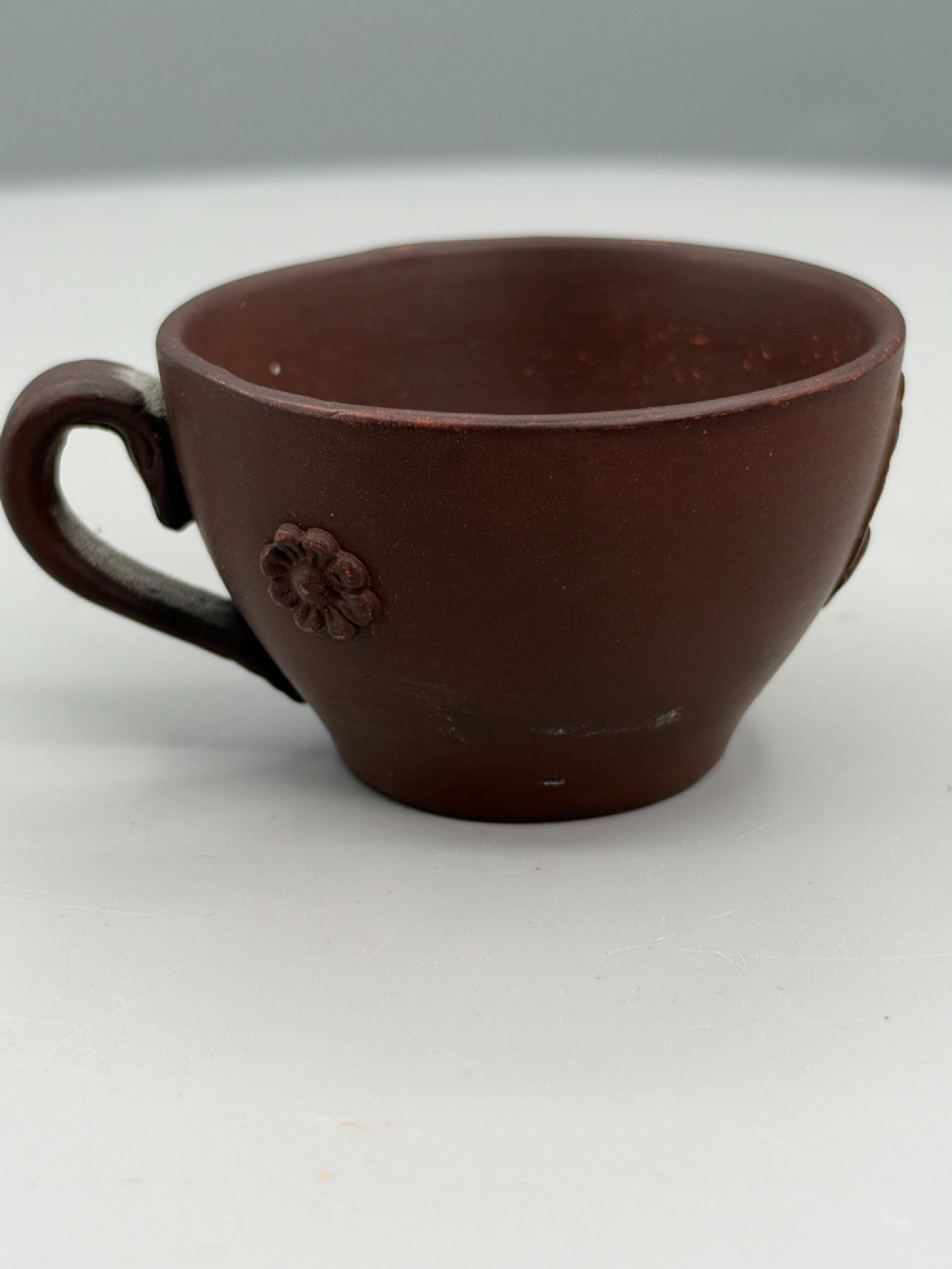 A small reddish-brown Staffordshire red stoneware teacup featuring a loop handle and an applied raised flowerhead sprig motif on its exterior.