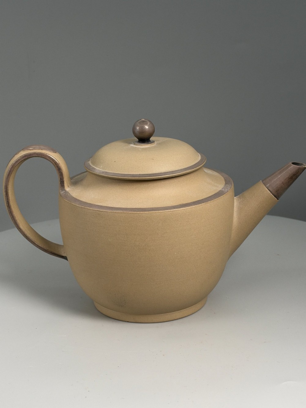 An 18th-century unglazed buff stoneware teapot with dark brown rim bands, a curved handle, and a round lid finial.