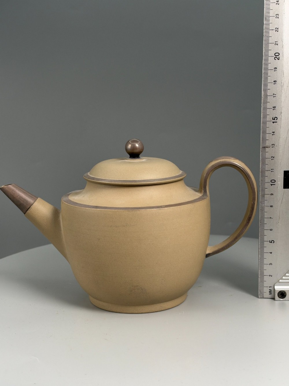 An 18th-century unglazed buff stoneware teapot with dark brown rim bands, a curved handle, and a round lid finial. View II