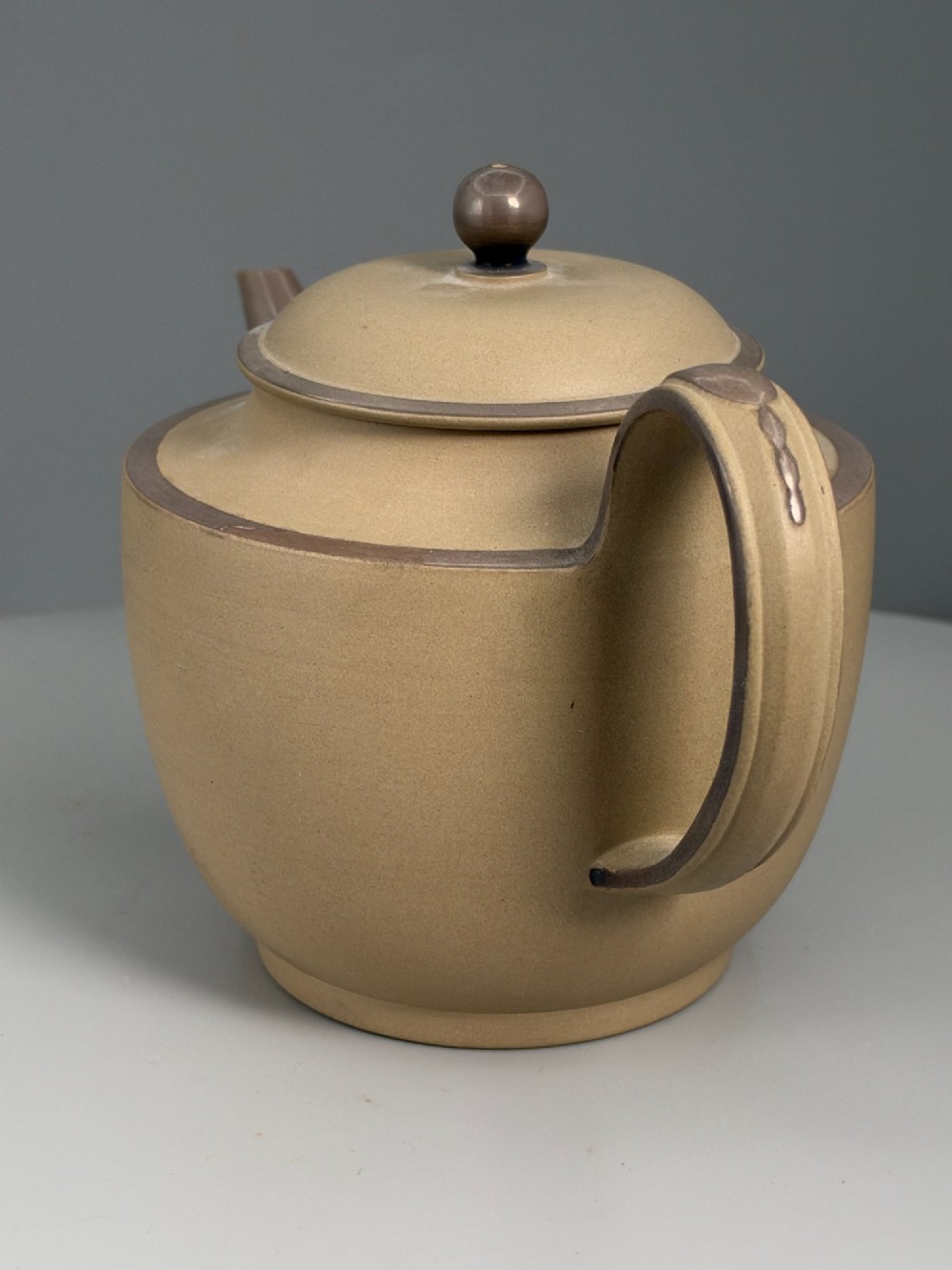 An 18th-century unglazed buff stoneware teapot with dark brown rim bands, a curved handle, and a round lid finial. View V
