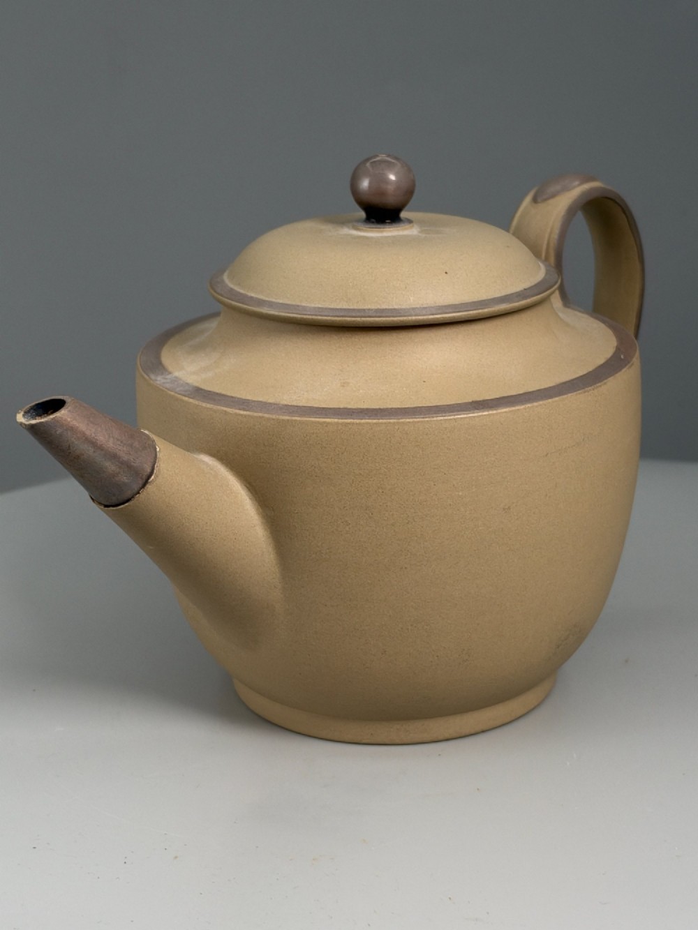 An 18th-century unglazed buff stoneware teapot with dark brown rim bands, a curved handle, and a round lid finial. View VI