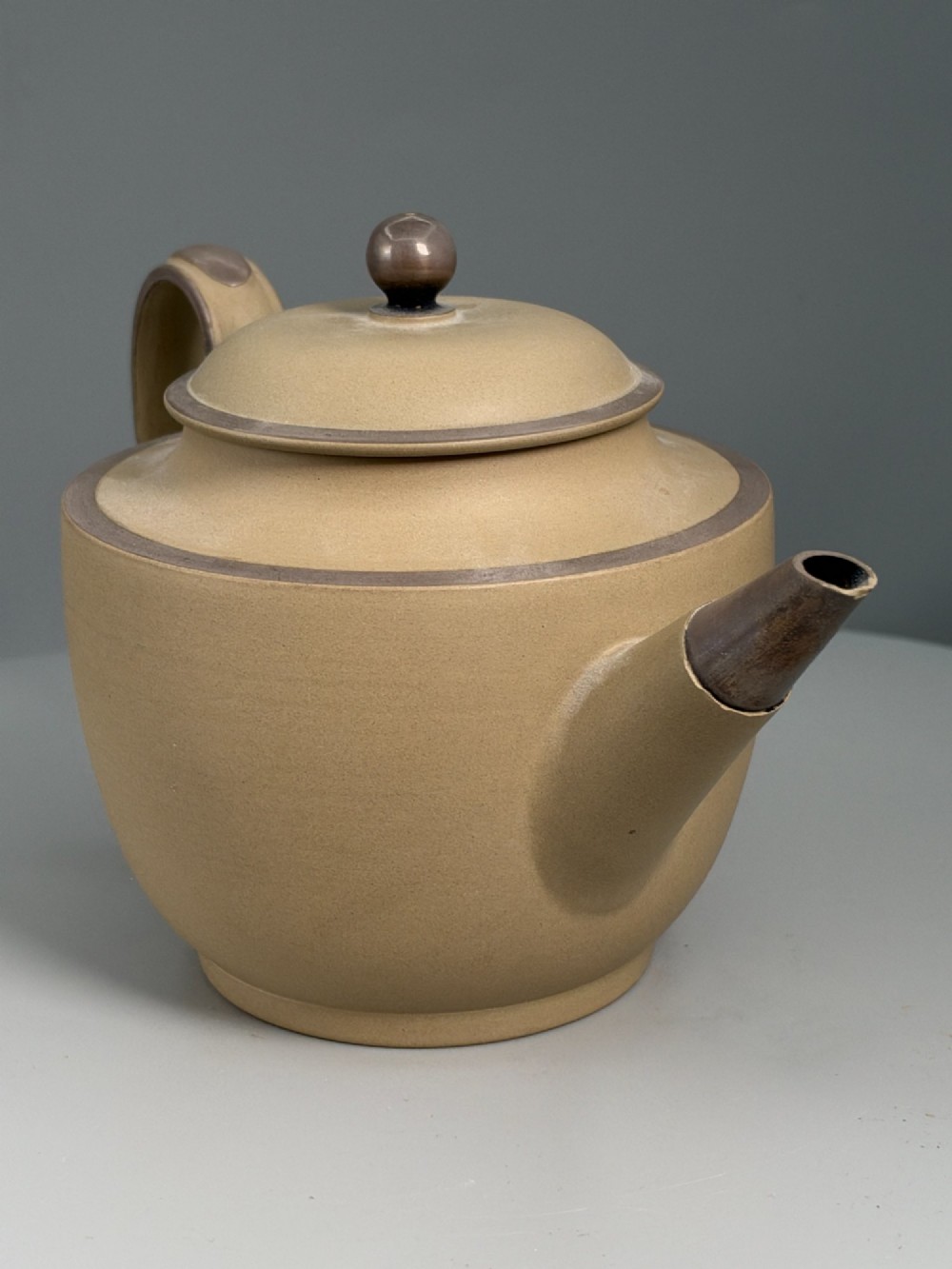 An 18th-century unglazed buff stoneware teapot with dark brown rim bands, a curved handle, and a round lid finial. View VII