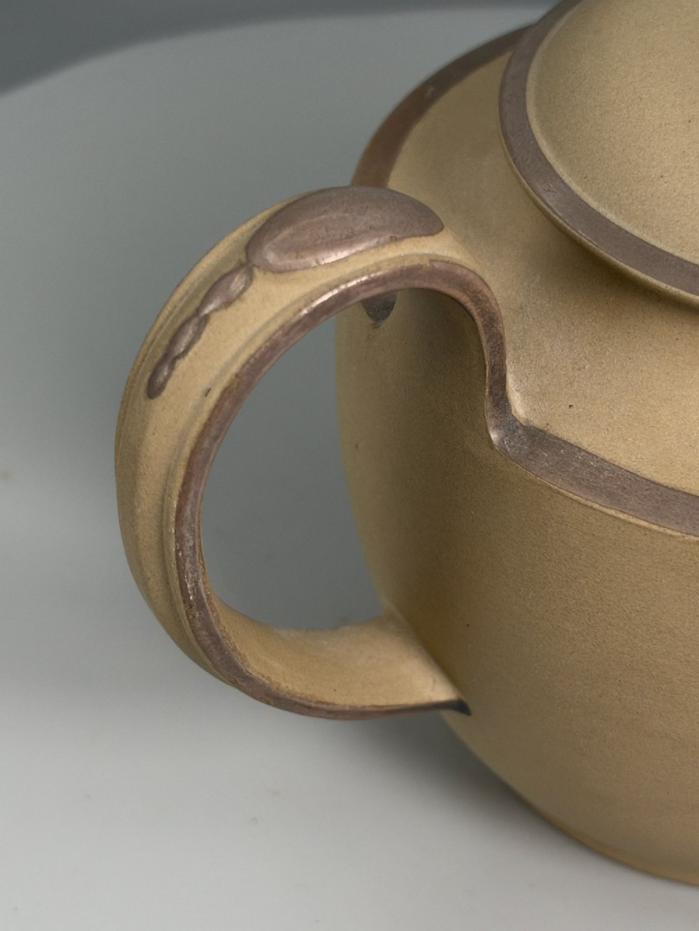 An 18th-century unglazed buff stoneware teapot with dark brown rim bands, a curved handle, and a round lid finial. View IX