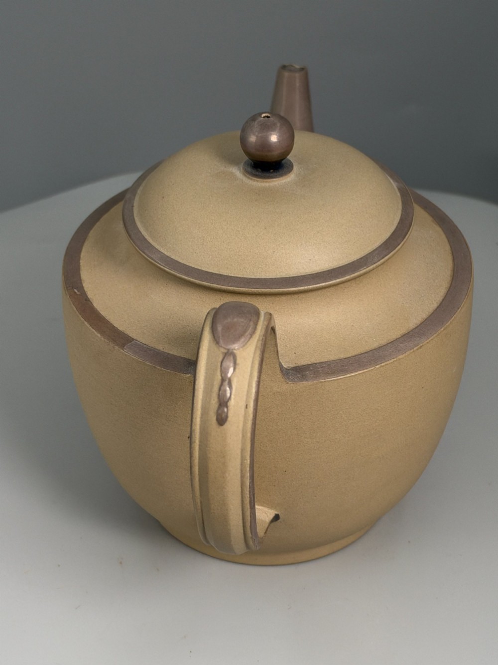 An 18th-century unglazed buff stoneware teapot with dark brown rim bands, a curved handle, and a round lid finial. View XIV