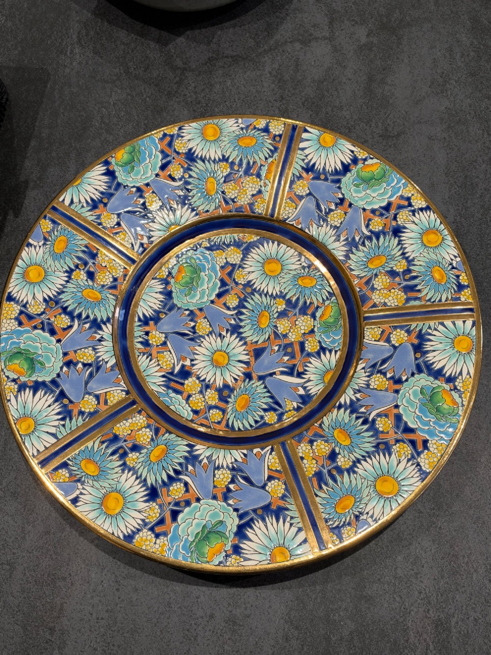 A circular Art Deco ceramic charger featuring radial segmented panels decorated with blue, turquoise, yellow, and white floral enamel patterns bordered in gold.