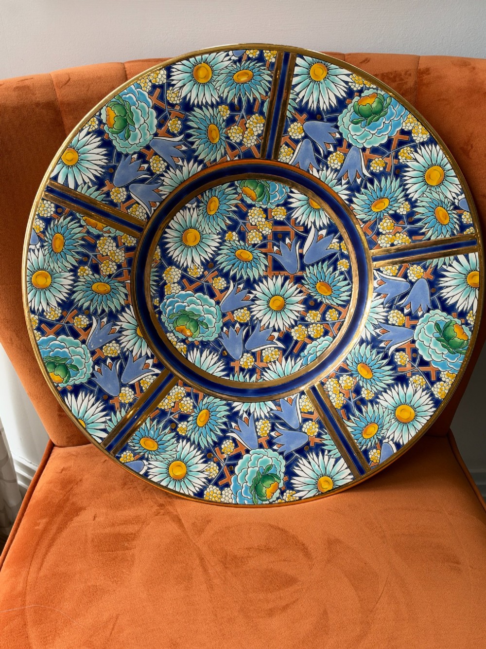 A circular Art Deco ceramic charger featuring radial segmented panels decorated with blue, turquoise, yellow, and white floral enamel patterns bordered in gold. View III