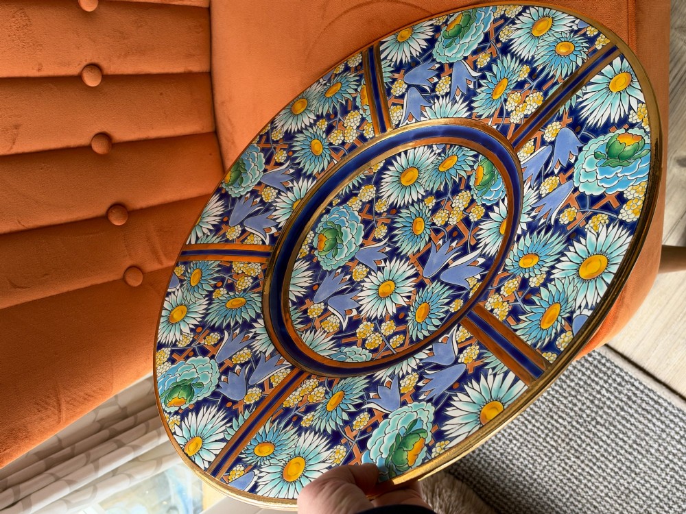 A circular Art Deco ceramic charger featuring radial segmented panels decorated with blue, turquoise, yellow, and white floral enamel patterns bordered in gold. View IV