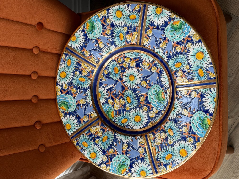A circular Art Deco ceramic charger featuring radial segmented panels decorated with blue, turquoise, yellow, and white floral enamel patterns bordered in gold. View V