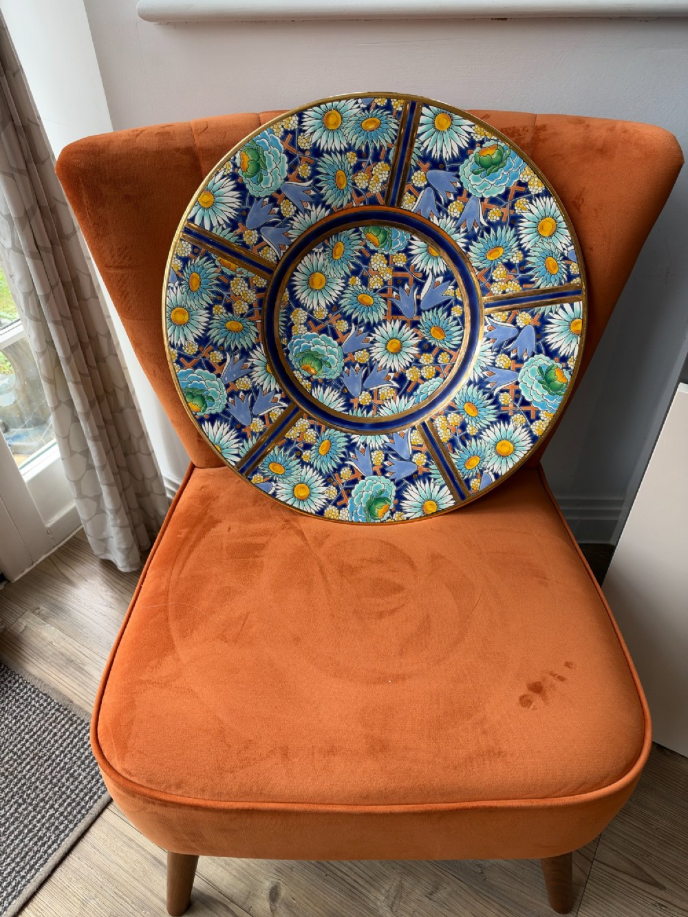A circular Art Deco ceramic charger featuring radial segmented panels decorated with blue, turquoise, yellow, and white floral enamel patterns bordered in gold. View VI