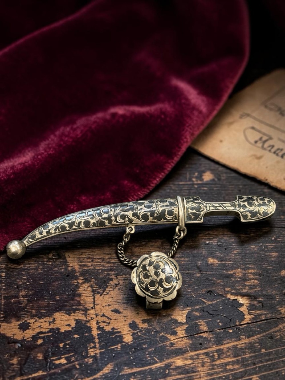 An antique Caucasian curved silver miniature dagger brooch featuring black niello scrollwork motifs and an attached suspension chain with a matching circular clasp.