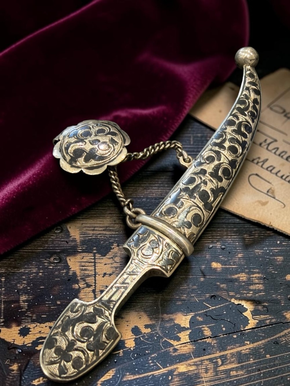 An antique Caucasian curved silver miniature dagger brooch featuring black niello scrollwork motifs and an attached suspension chain with a matching circular clasp. View III