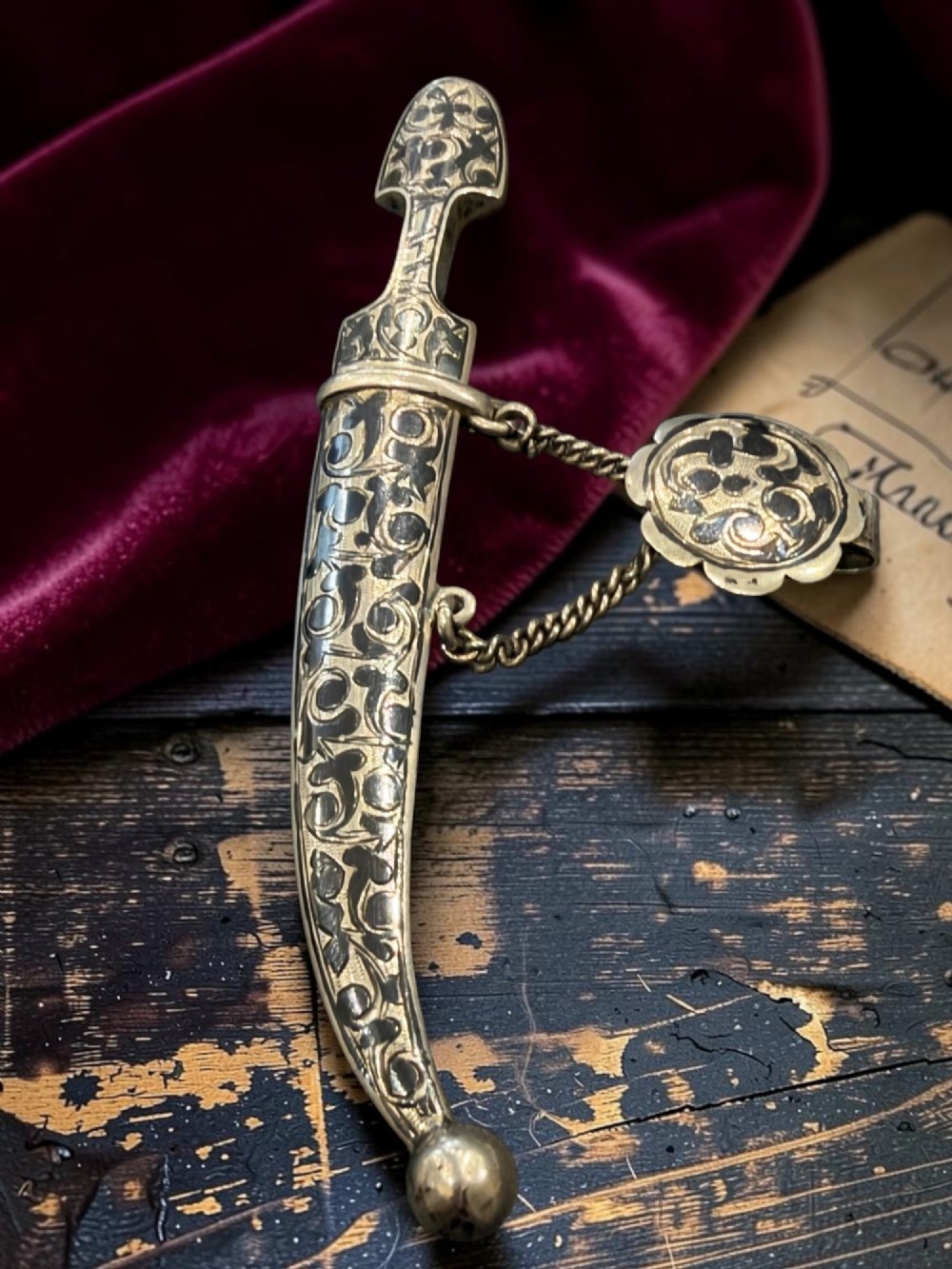 An antique Caucasian curved silver miniature dagger brooch featuring black niello scrollwork motifs and an attached suspension chain with a matching circular clasp. View IV