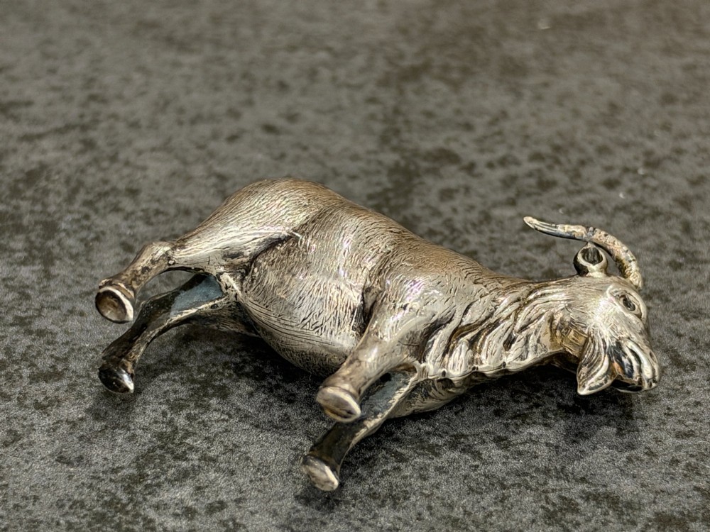 A small cast sterling silver miniature figurine of a billy goat displaying detailed fur texture, curved horns, and British hallmarks stamped along its side. View IX