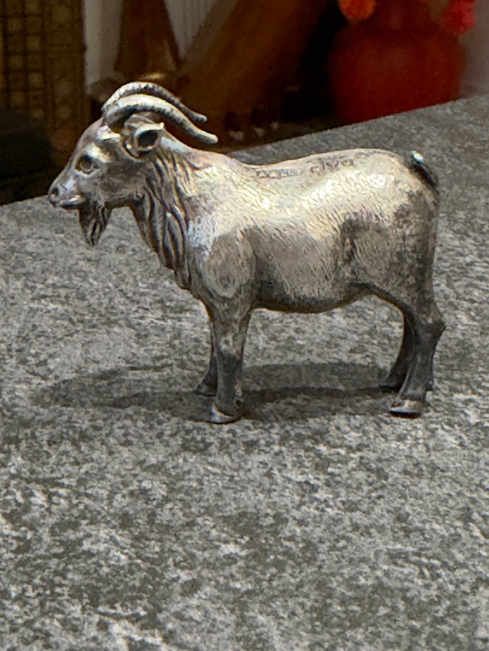 A small cast sterling silver miniature figurine of a billy goat displaying detailed fur texture, curved horns, and British hallmarks stamped along its side. View XIV