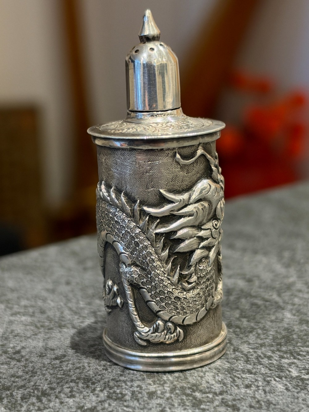 A cylindrical silver pepper shaker decorated with a detailed dragon repousse motif against a textured background with a pointed finial cap. View III