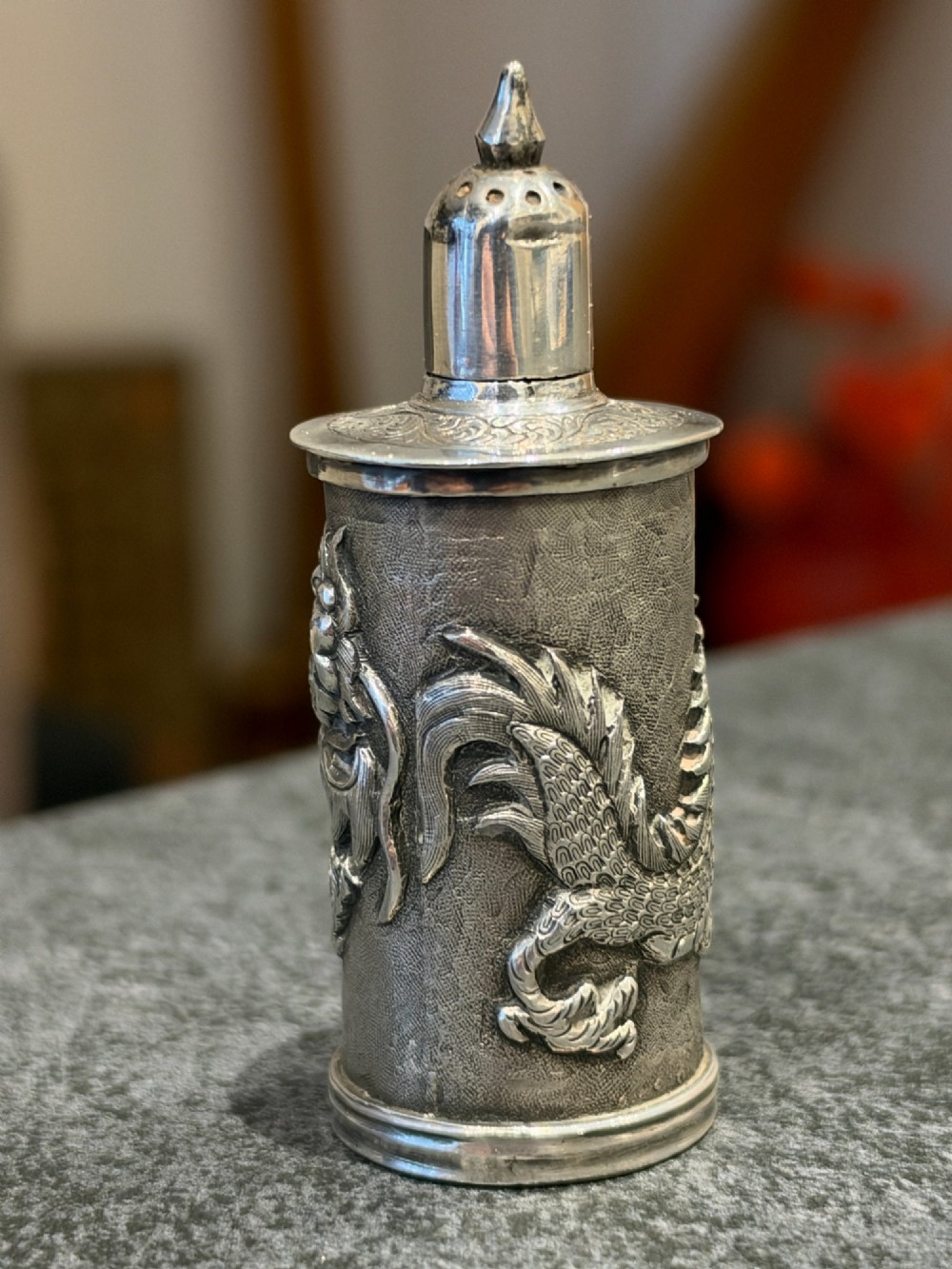 A cylindrical silver pepper shaker decorated with a detailed dragon repousse motif against a textured background with a pointed finial cap. View V