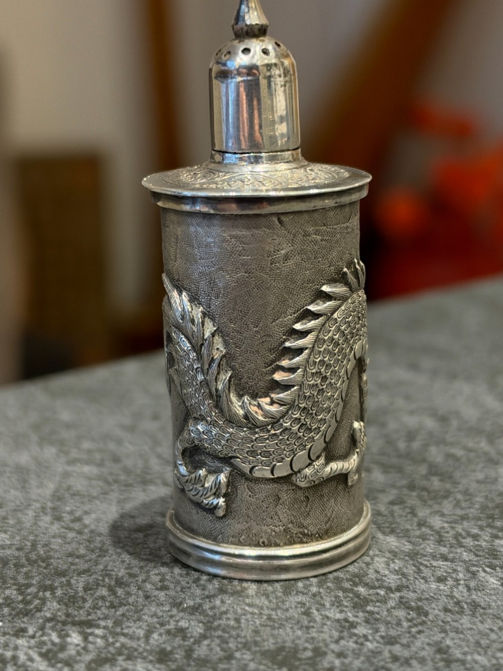 A cylindrical silver pepper shaker decorated with a detailed dragon repousse motif against a textured background with a pointed finial cap. View VI