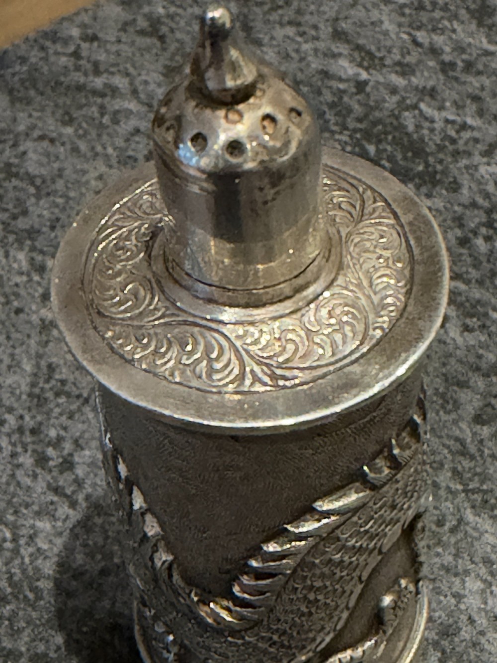 A cylindrical silver pepper shaker decorated with a detailed dragon repousse motif against a textured background with a pointed finial cap. View X