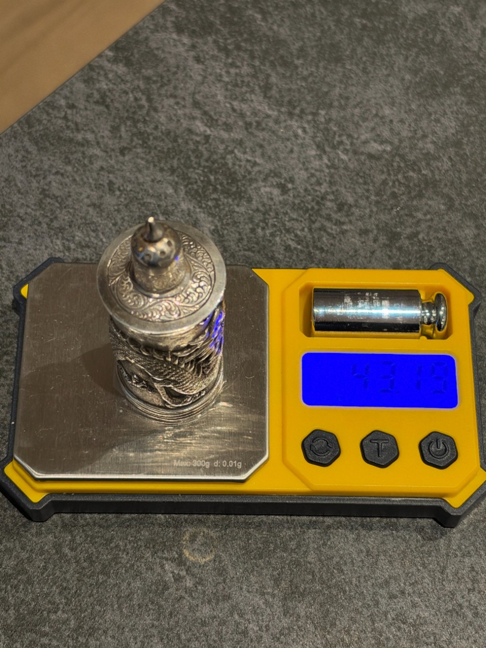 A cylindrical silver pepper shaker decorated with a detailed dragon repousse motif against a textured background with a pointed finial cap. View XII