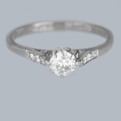 fig 1. An 18ct white gold solitaire ring showcasing a central round diamond in a claw setting flanked by small diamonds in the shoulders.