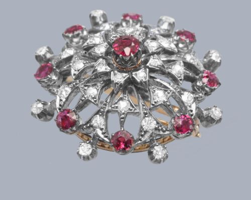fig 4. A circular Victorian brooch rendered in silver and gold, featuring a central round ruby surrounded by intricate diamond-set starburst openwork and an outer border of alternating rubies and diamonds.