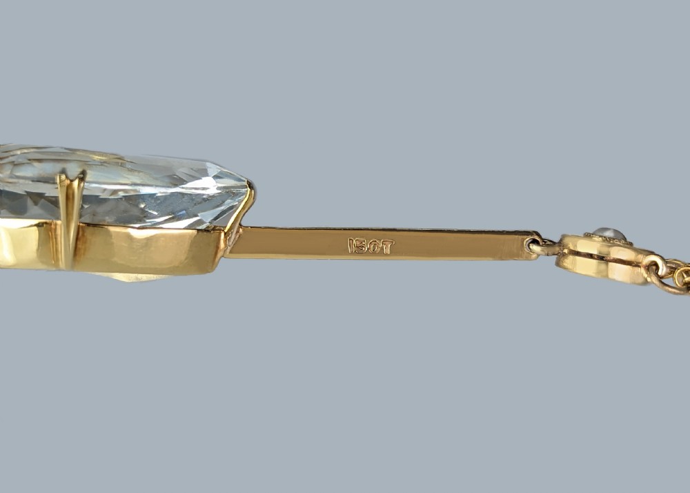 An Edwardian 15ct gold pendant featuring a circular accent set with a seed pearl, suspending a linear bar and pear-cut aquamarine gemstone on a fine gold chain. View X