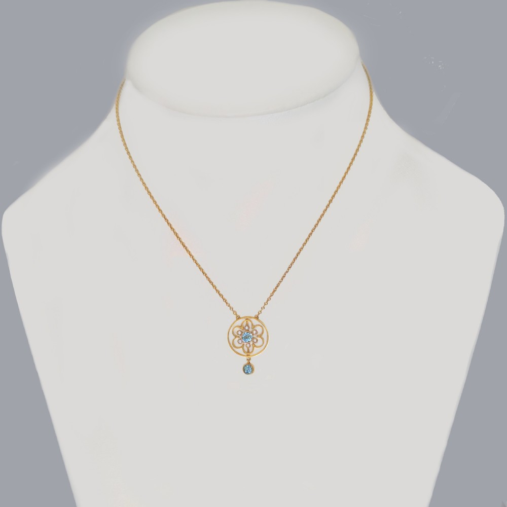An Edwardian 15ct gold openwork circular pendant featuring a central round aquamarine surrounded by seed pearls and a hanging aquamarine drop on a gold chain. View II