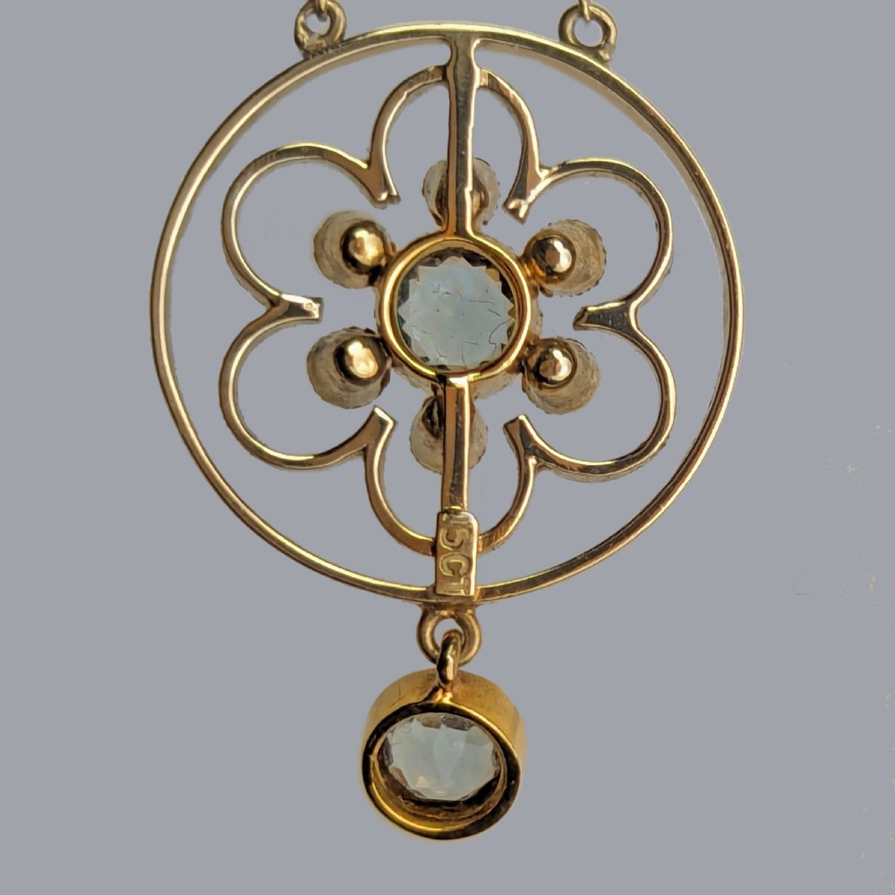 An Edwardian 15ct gold openwork circular pendant featuring a central round aquamarine surrounded by seed pearls and a hanging aquamarine drop on a gold chain. View V