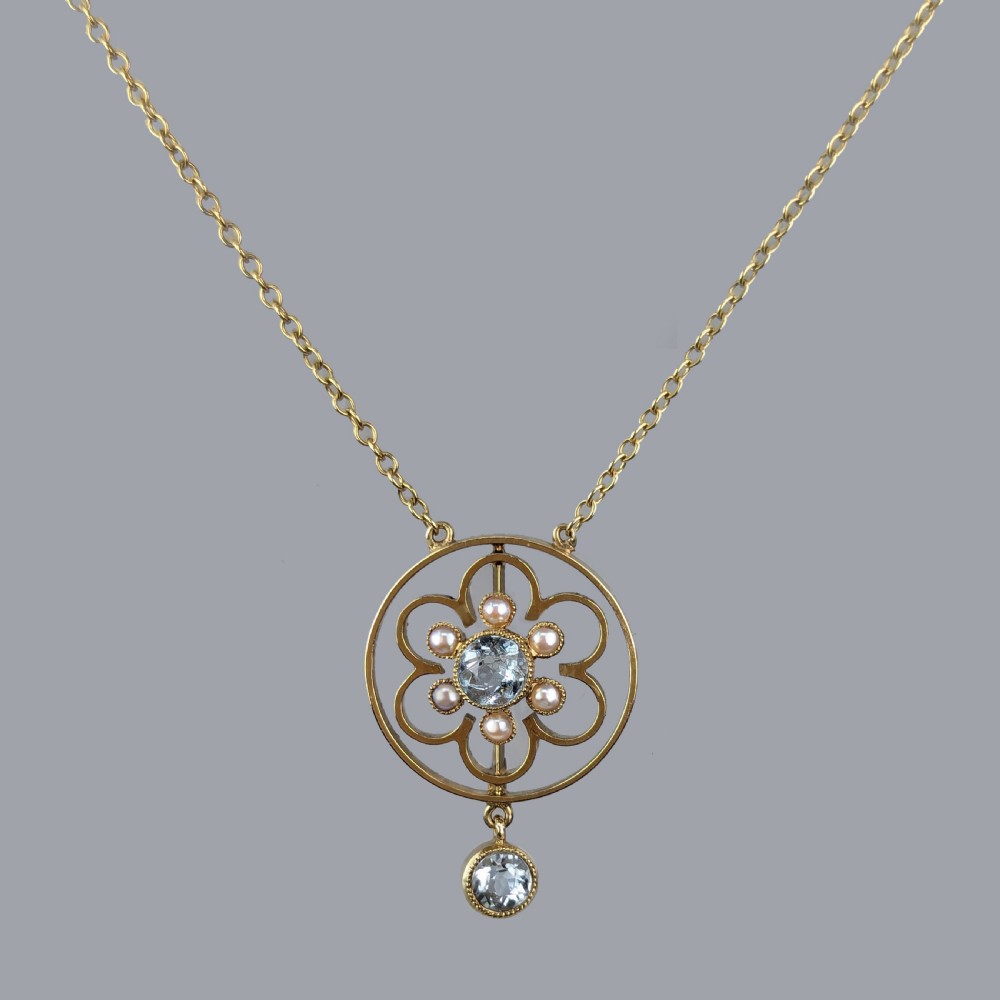 An Edwardian 15ct gold openwork circular pendant featuring a central round aquamarine surrounded by seed pearls and a hanging aquamarine drop on a gold chain. View VI