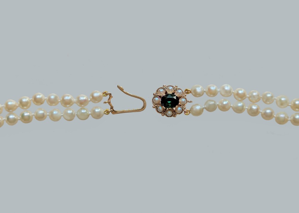 A double strand cream pearl necklace featuring an ornate gold clasp set with a central round green tourmaline stone. View VIII