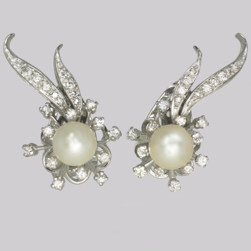 A pair of 1950s 18ct white gold clip-on earrings featuring central cultured pearls surrounded by diamond-set floral sprays and sweeping curved accents.