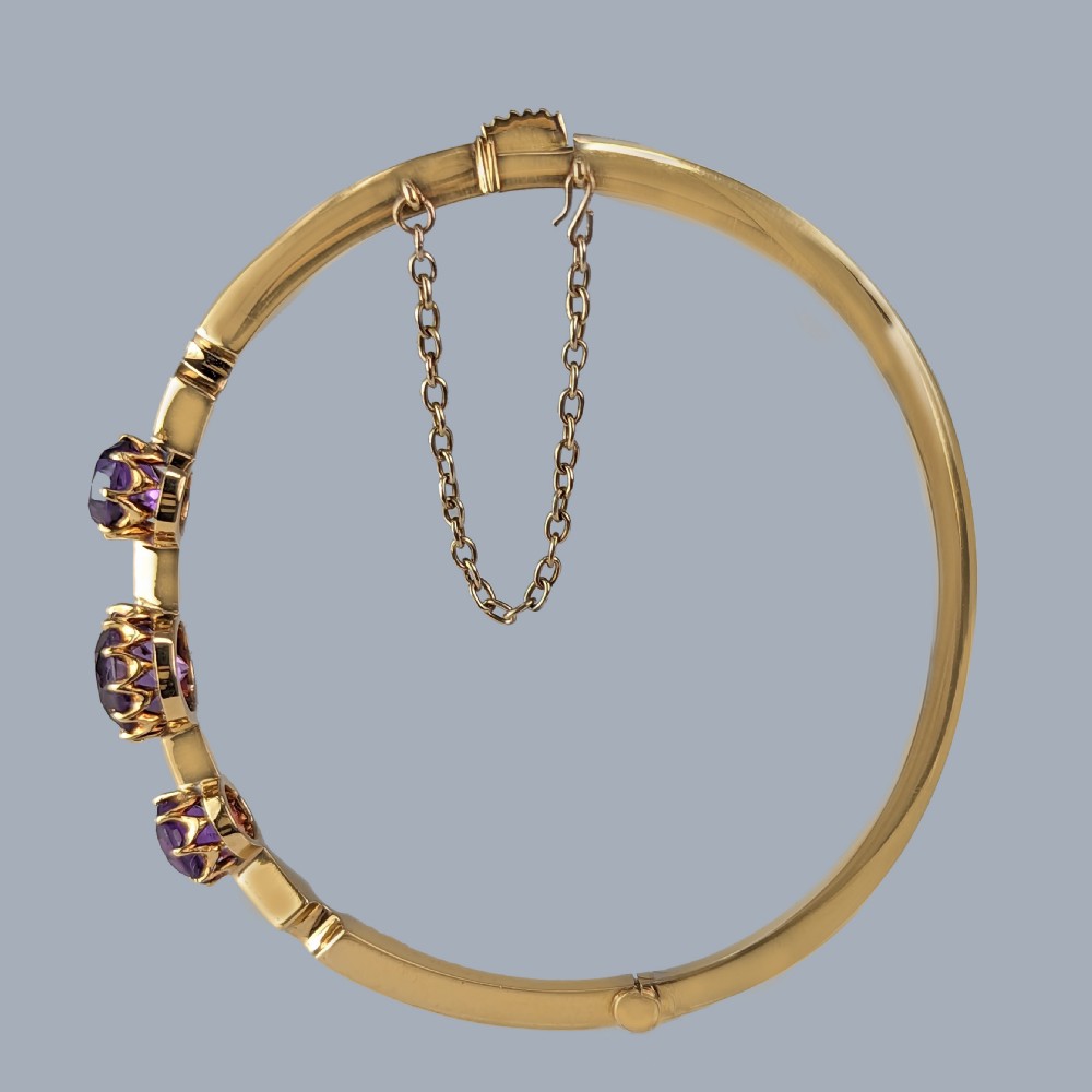 An 18ct yellow gold Victorian hinged bangle featuring three claw-set round purple amethysts and an attached safety chain. View VIII