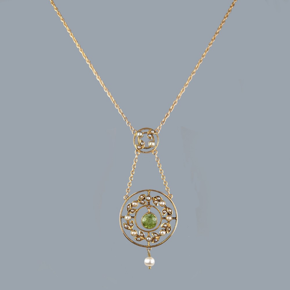 An Edwardian 15ct gold chain displaying a circular openwork garland pendant set with a central round green peridot surrounded by seed pearl leaves and a single pearl drop. View VII